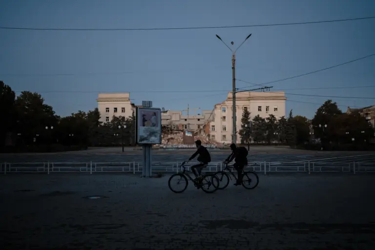 Bewohner fahren eine Straße gegenüber der durch Beschuss zerstörten Regionalverwaltung entlang. Cherson, Region Taurien. Projekt „Frontnahe Städte“. Oktober 2025. Foto: Wjatscheslaw Ratynskyj.