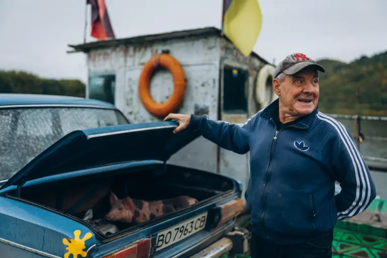 Muž přepravuje sele přes Dnistr na trajektu a ukládá ho do kufru svého auta. Na hranici Bukoviny a Podillja. Říjen 2025. Expedice Podilljím. Foto: Andrij Jakymenko.